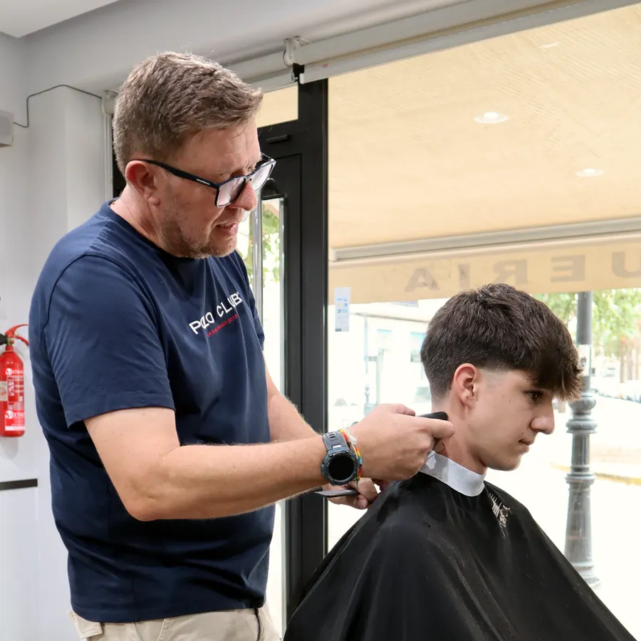 El barbero de Rapiño cortando el pelo a un cliente joven junto al escaparate de la peluquería.